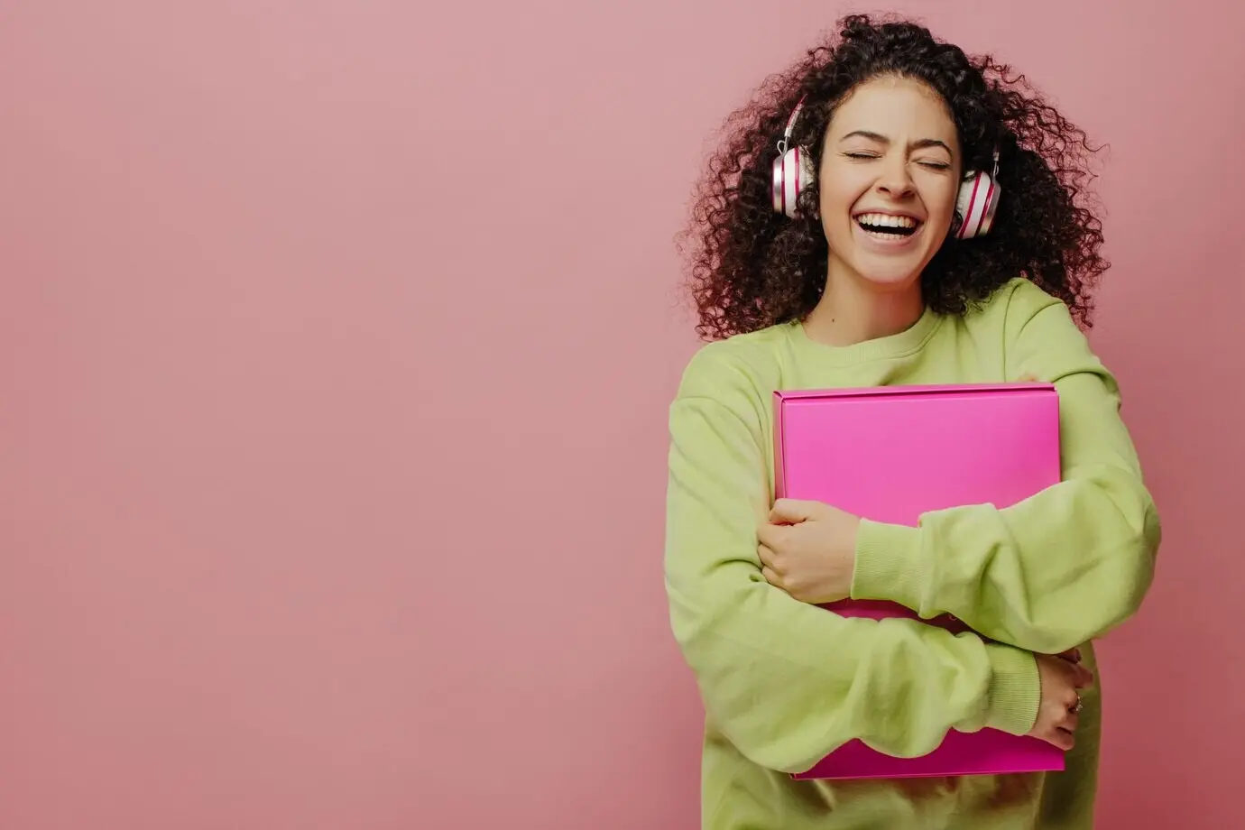 Ein junges, hellhäutiges, lockiges, brünettes Mädchen lacht mit geschlossenen Augen und umklammert eine Schachtel an der Brust, vor einem Hintergrund mit Platz für Text. Mit Kopfhörern hört sie ihre Lieblingsmusik – Konzept des Genießens des Moments.